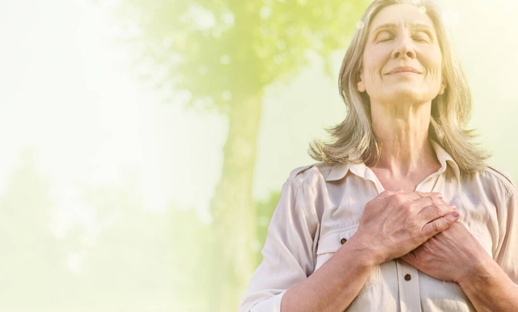 A mature woman stands outdoors with her hands over her chest, expressing serenity against a softly lit green background.